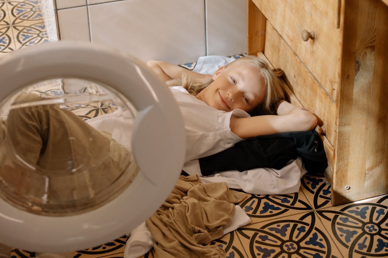 creative-02 A smiling young girl lies on a tiled floor surrounded by laundry indoors.