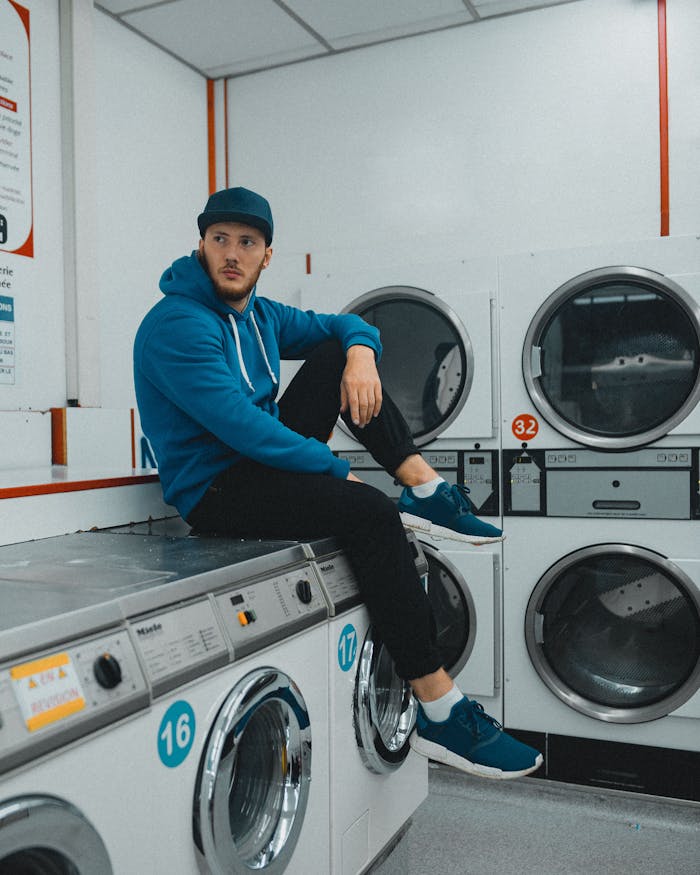 mobile-02 A man casually sitting in a laundromat, wearing a hoodie and cap, with washing machines in view.